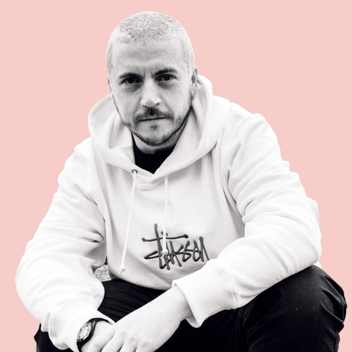 Man with a short haircut and beard, wearing a white hoodie and dark pants, sitting against a soft pink background. He has a thoughtful expression.