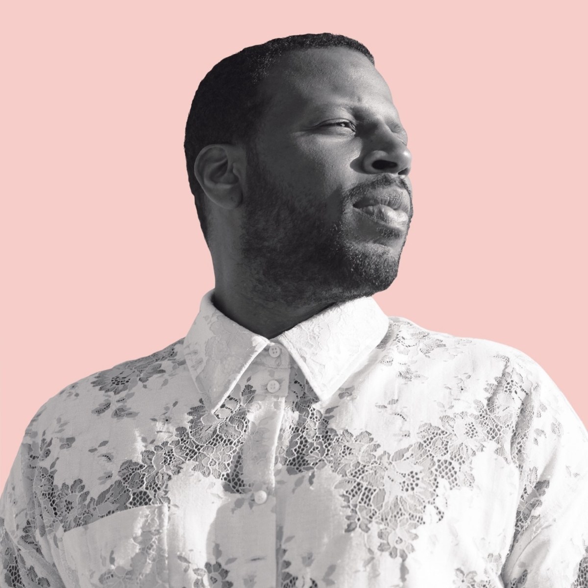 A man gazes sideways with a thoughtful expression. He wears a white lace shirt, standing against a plain pink background, conveying a serene mood.