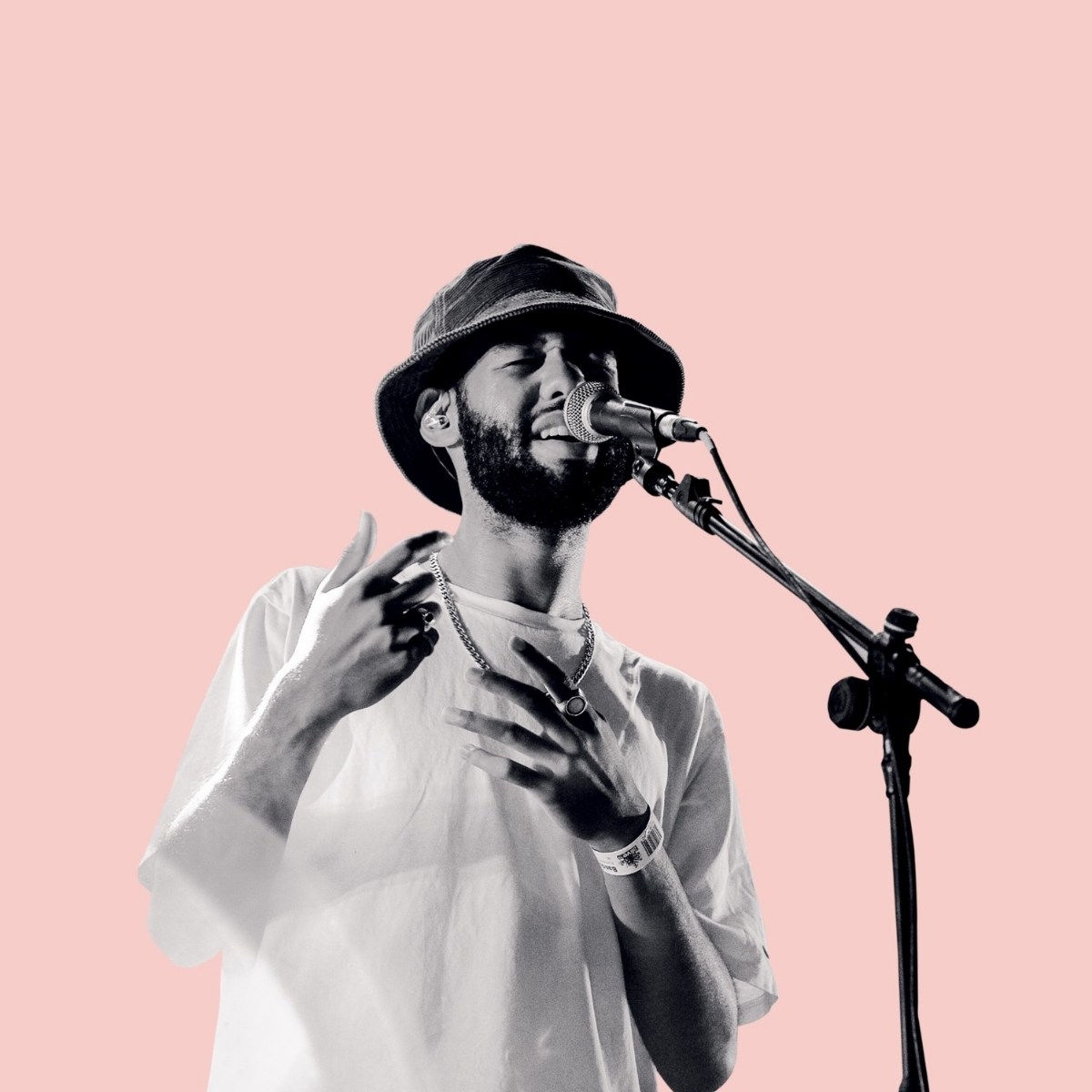A man wearing a bucket hat passionately sings into a microphone against a pink background. He gestures expressively, conveying emotion and energy.