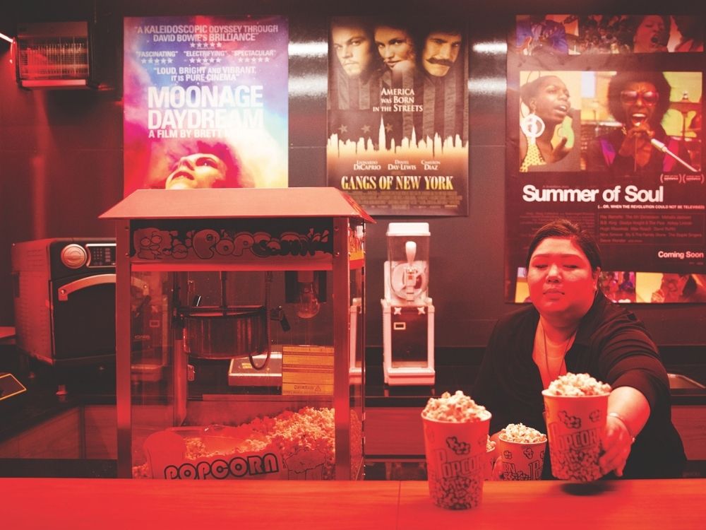 A person serves popcorn in a dimly lit movie theater concession stand. Behind, colorful posters for "Moonage Daydream," "Gangs of New York," and "Summer of Soul" are displayed. The scene conveys a nostalgic, cinematic atmosphere.