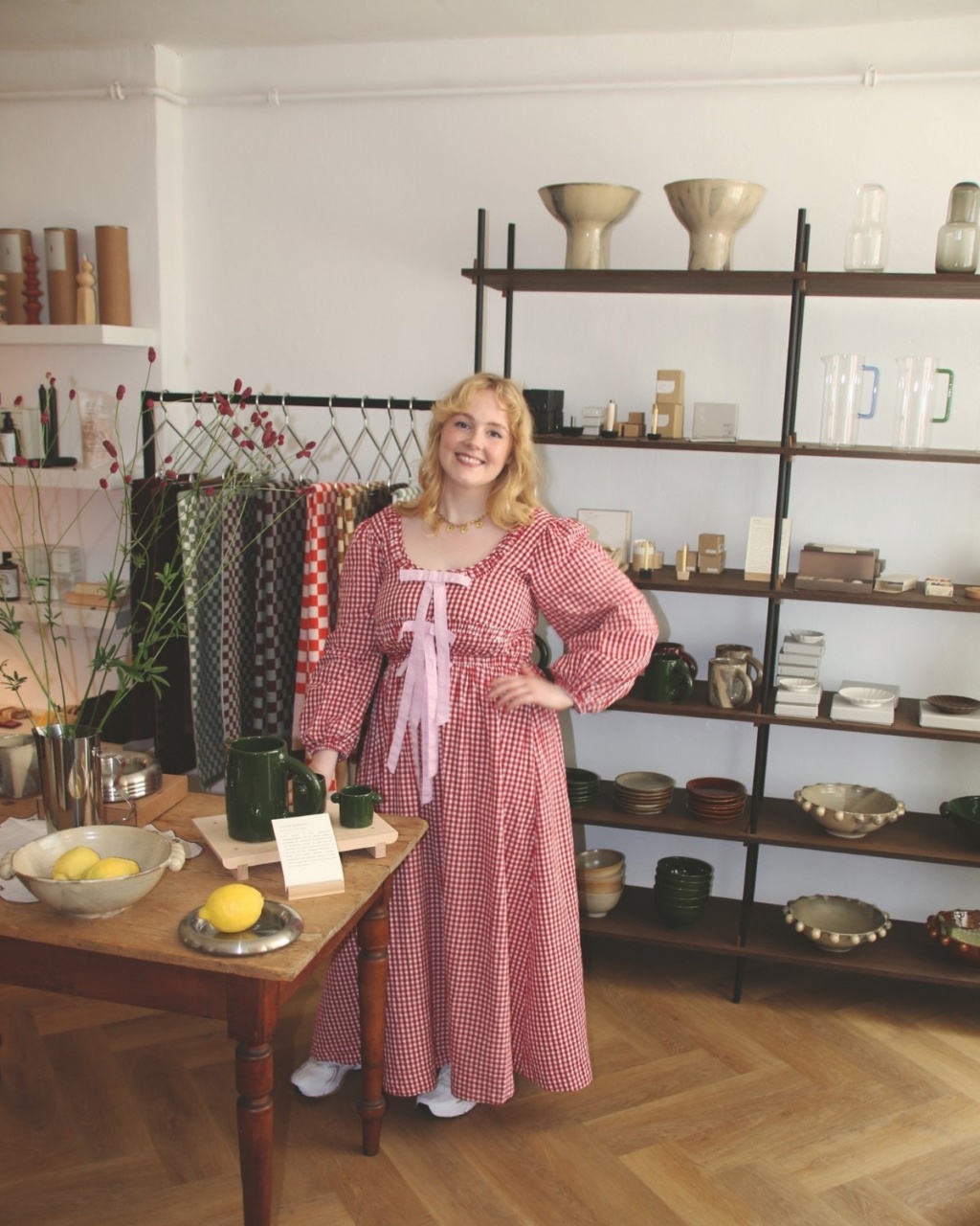 A person in a red checkered dress stands smiling in a cozy shop. The room features rustic pottery, textiles, and a wooden table with lemons. The mood is warm and inviting.