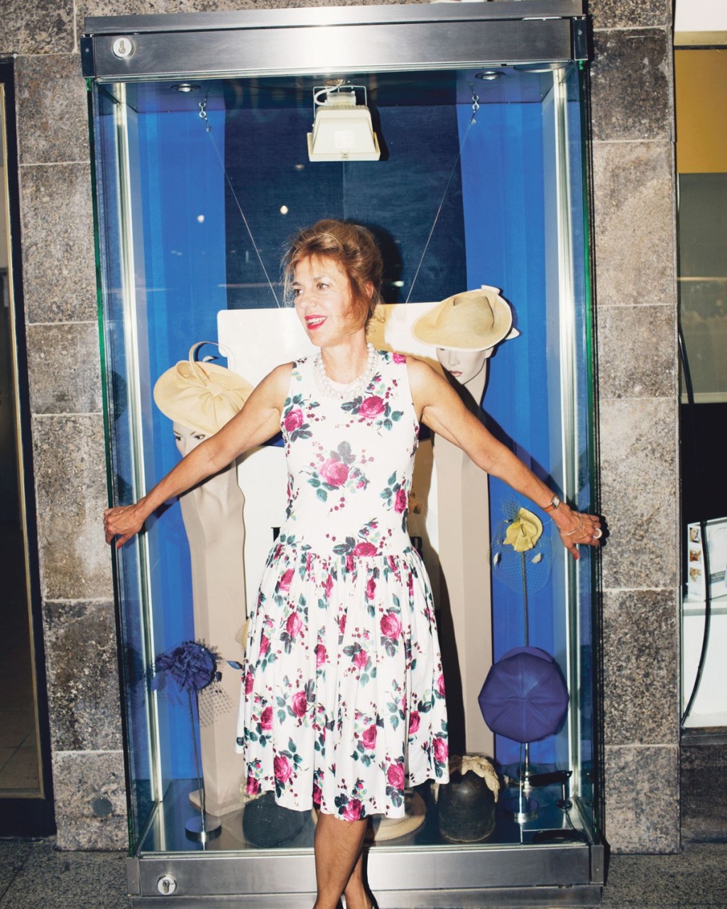 A woman poses in a shop window wearing a floral dress, surrounded by mannequins and various props.