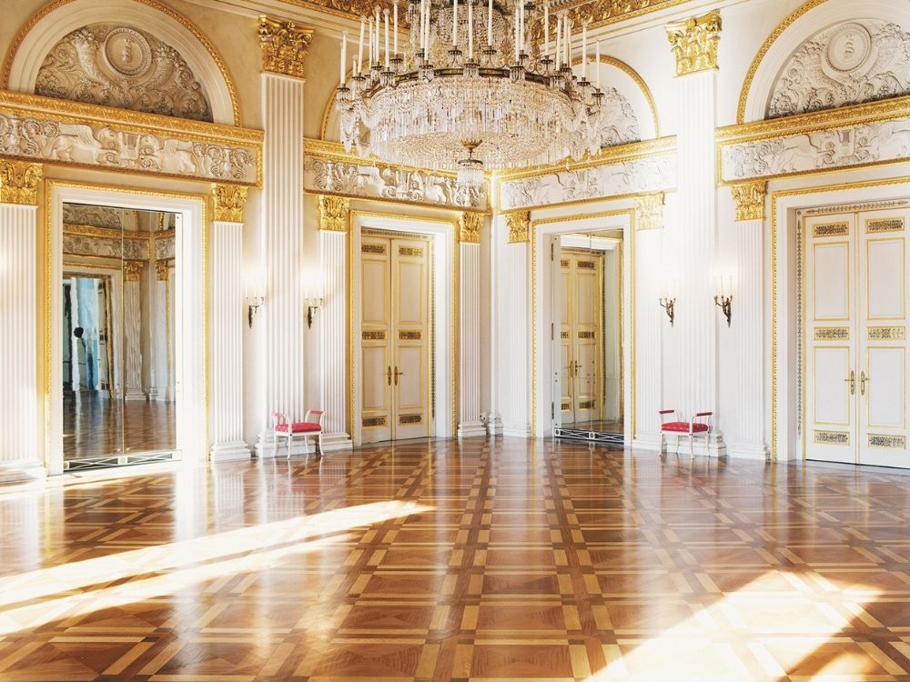 Luxurious ballroom with ornate gold detailing, high ceilings, and a grand chandelier. Elegant parquet flooring reflects natural light, conveying opulence and grandeur.