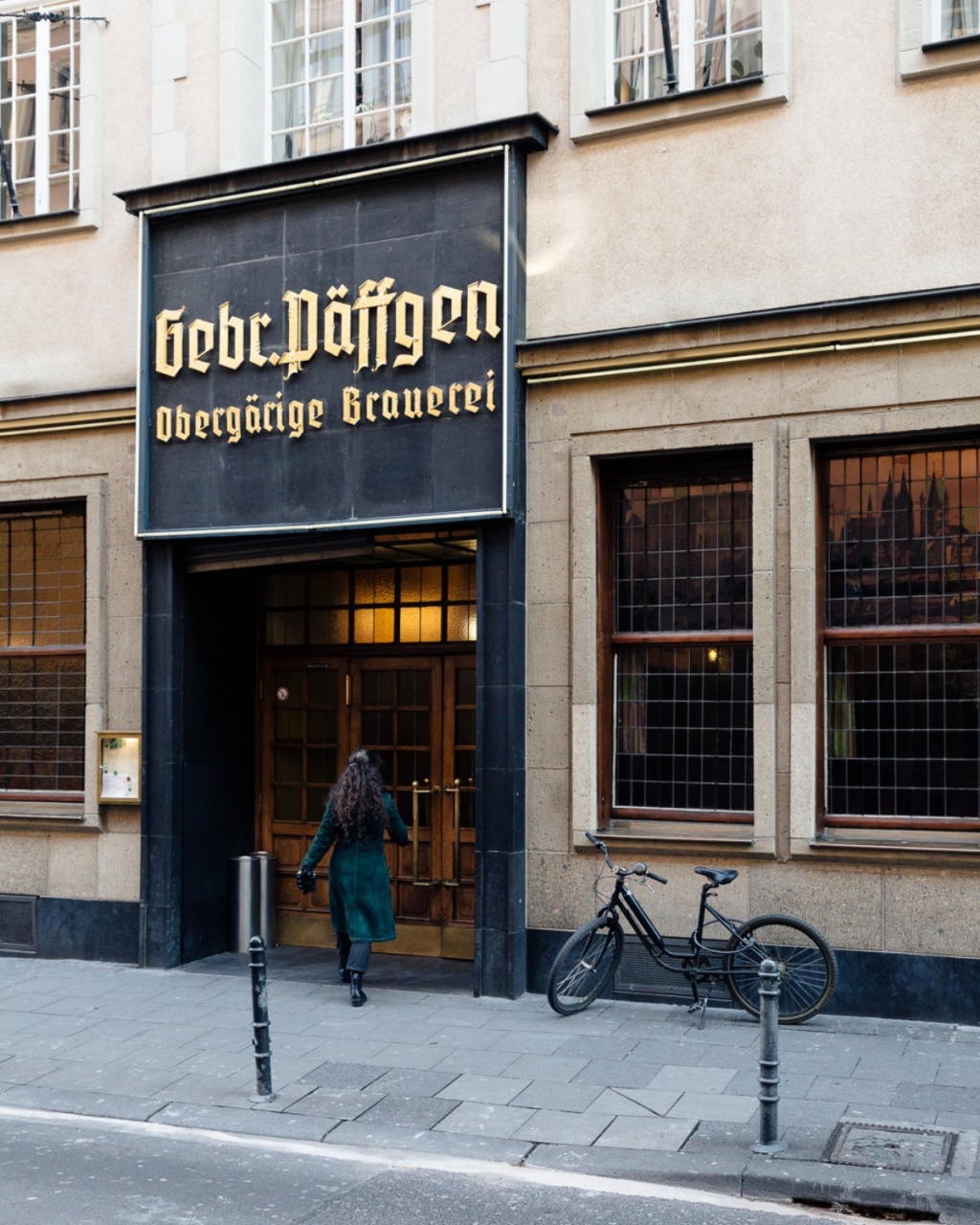 A person in a long green coat walks into a historic brewery with a large black sign reading "Gebr. Päffgen Obergärige Brauerei." A bicycle is parked outside.