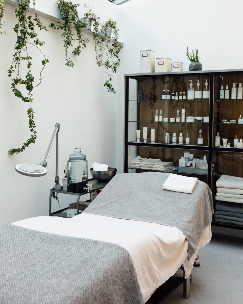 A serene spa room features a treatment bed with gray and white linens, surrounded by lush hanging plants. Glass cabinets display skincare products, evoking tranquility.