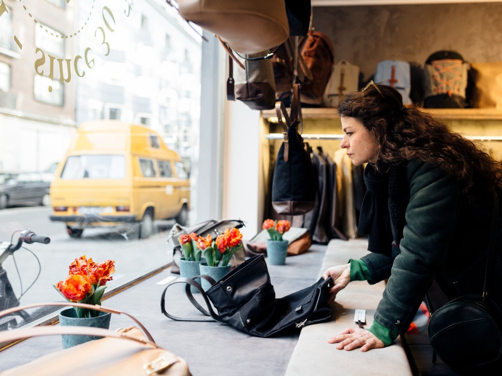 A woman in a green coat examines handbags in a store window. Outside, a yellow van and city buildings are visible. Tulips add a bright touch.
