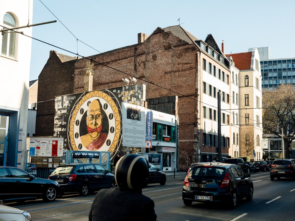 Street scene featuring a large mural on a building wall, depicting a person with a headscarf in a circular design. Cars and a motorcyclist in foreground.