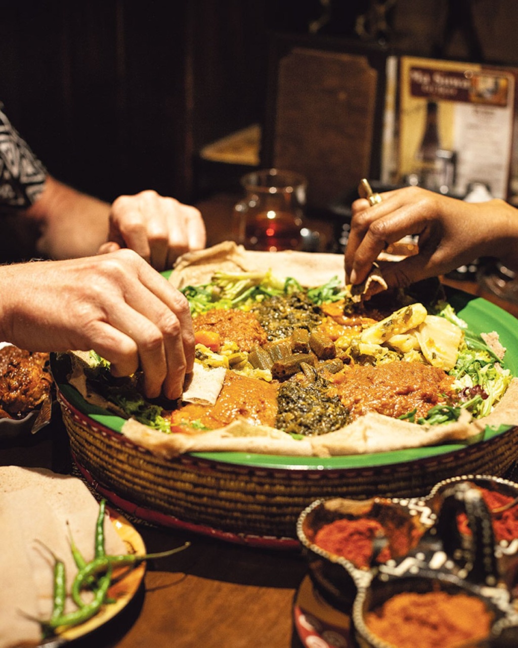 Hands of diverse diners reach for colorful Ethiopian dishes on a large shared platter. The atmosphere is warm and communal, conveying a sense of togetherness.