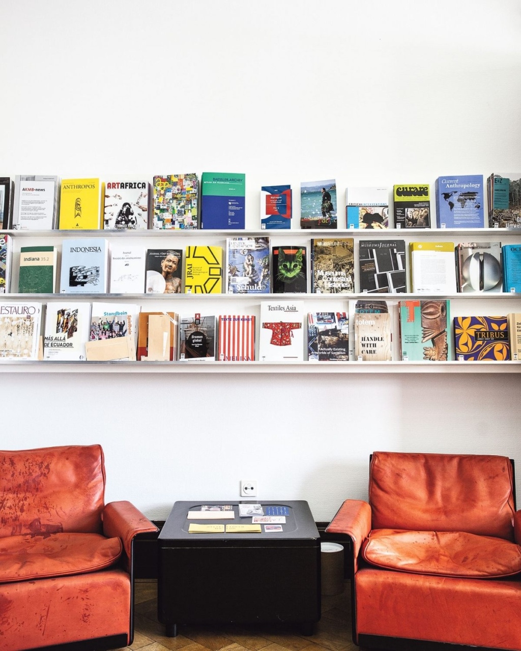 Two red leather chairs face a small black table in a cozy reading nook. Above, colorful books line three white shelves against a clean, white wall.