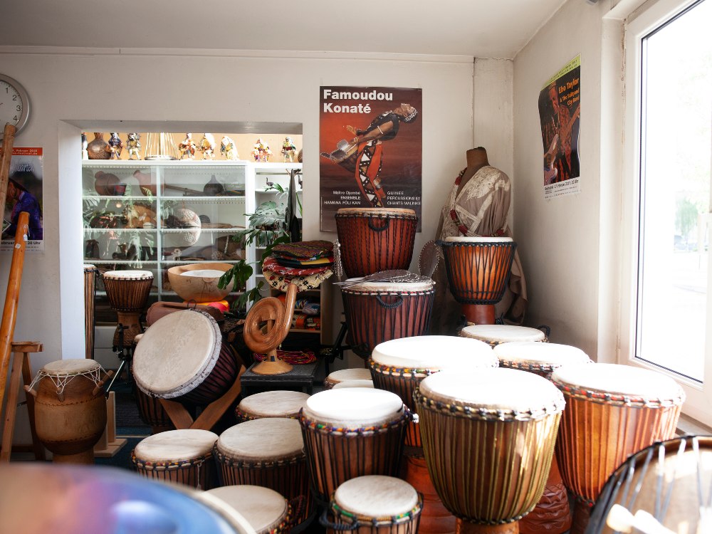 A room filled with various colorful hand drums, including djembes, arranged neatly. Posters on the wall; natural light filters in, creating a vibrant atmosphere.