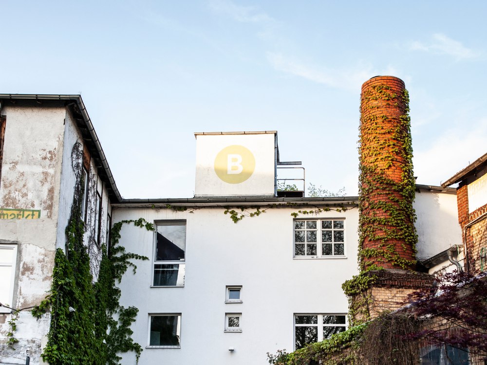 A white building with a tall, ivy-covered brick chimney and a yellow circle with a "B" on its roof. The setting is calm under a blue sky.