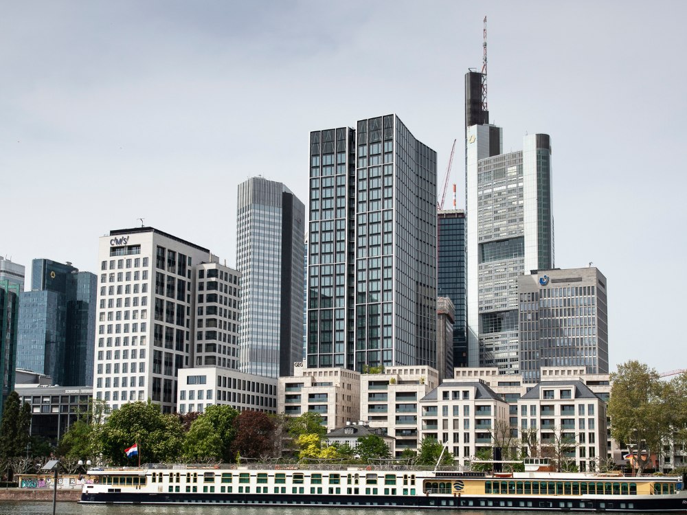 Skyline of modern skyscrapers and office buildings against a gray sky. A cruise ship with trees nearby is in the foreground, evoking an urban, bustling atmosphere.