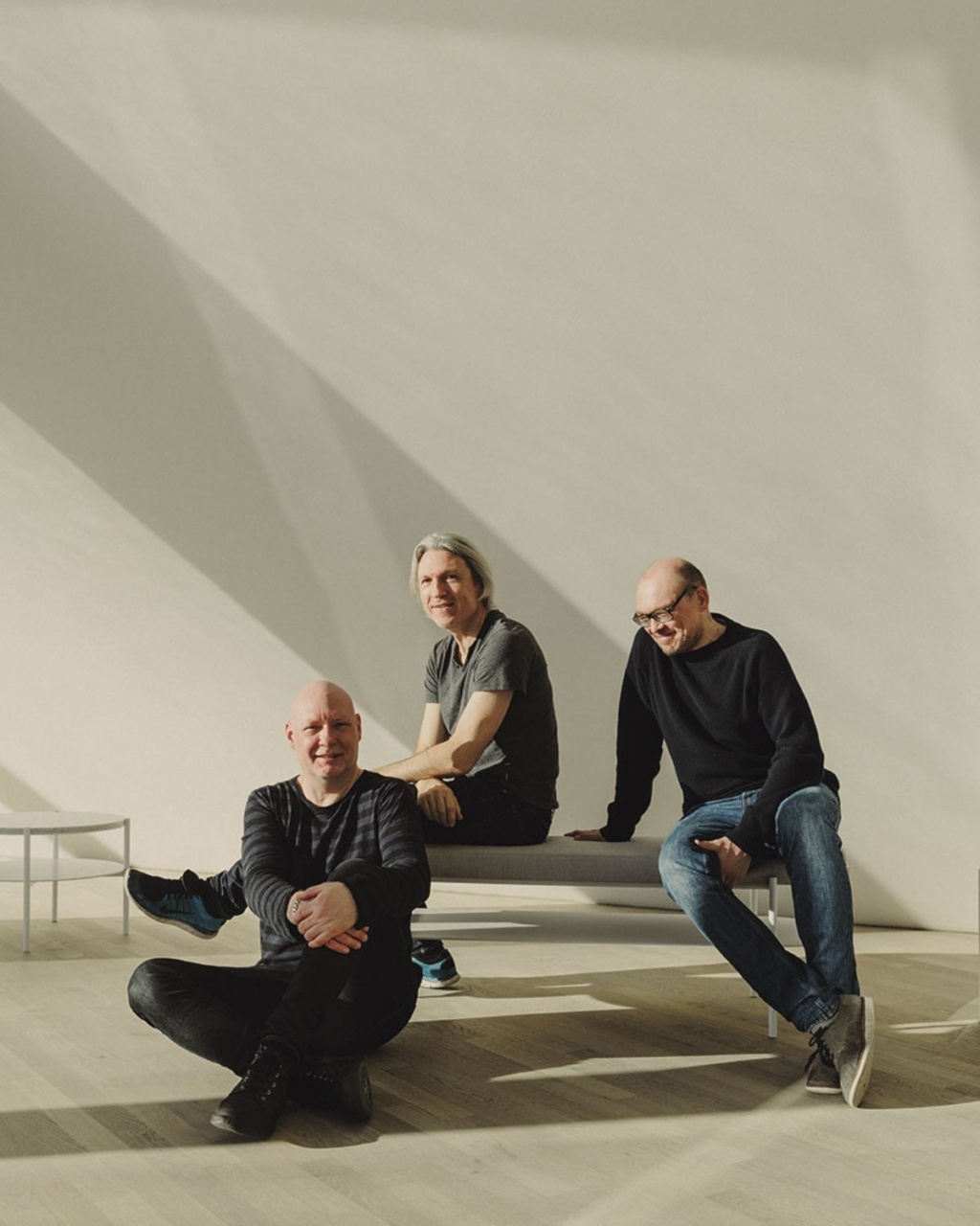 Three men are casually sitting and smiling in a sunlit room with light wood flooring. One sits cross-legged on the floor, the other two on a bench.