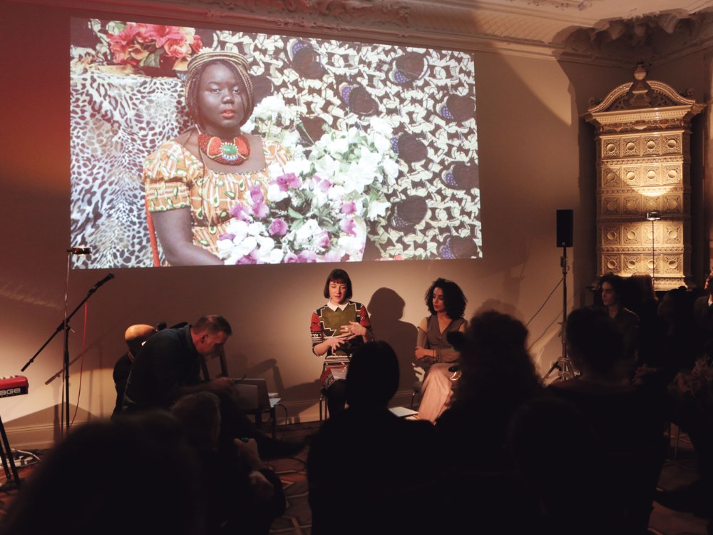 A woman in patterned attire holds flowers in a vivid photo projected on a wall behind a speaker and audience in a warmly lit room, creating a reflective mood.