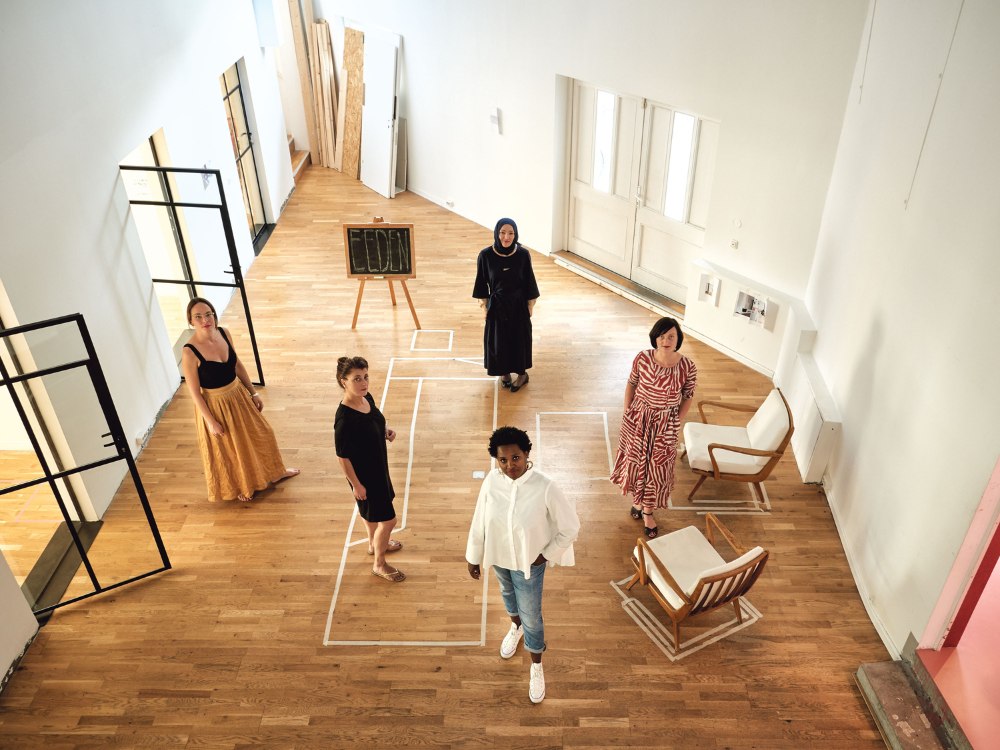A group of five diverse people stand on a wooden floor in a spacious room with white walls. A chalkboard labeled "Eeden" is in the background, set against a minimalistic decor with several chairs.
