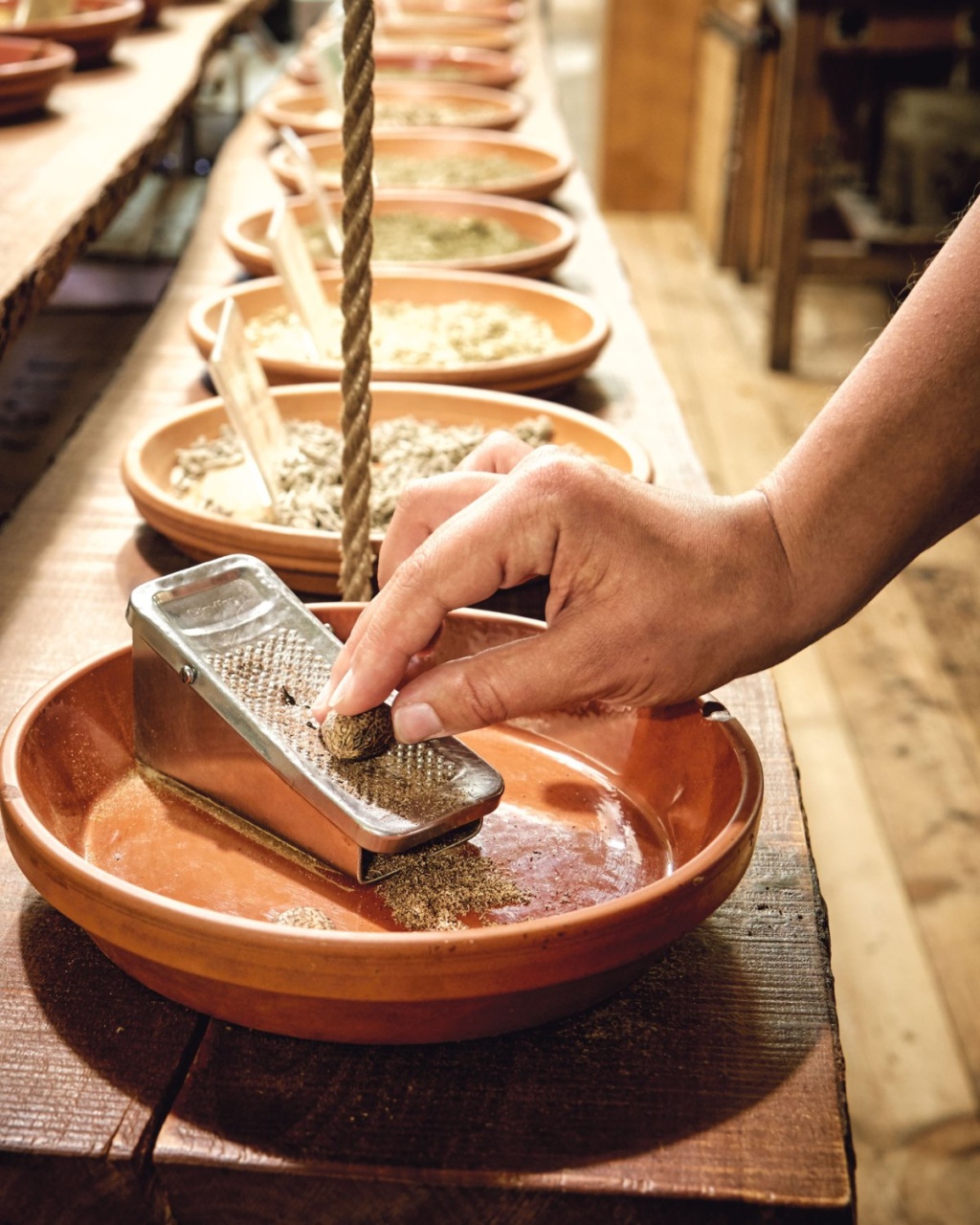 A hand grates a spice over a terracotta dish, surrounded by rows of similar bowls filled with spices on a wooden surface, giving a warm, rustic feel.