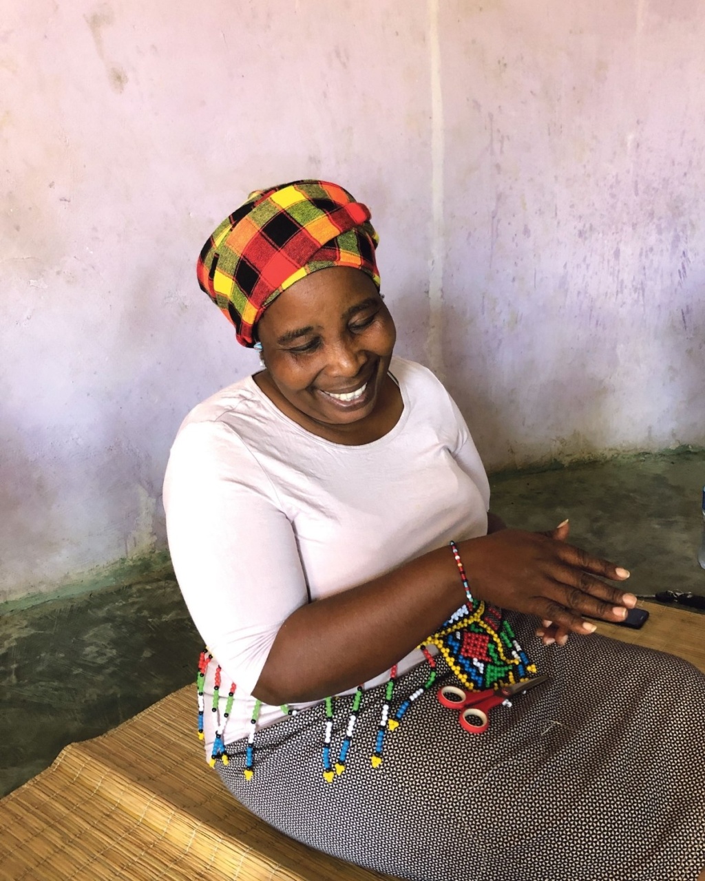 A woman sits indoors on a woven mat, smiling warmly. She wears a colorful plaid headscarf and beaded jewelry, conveying joy and cultural richness.