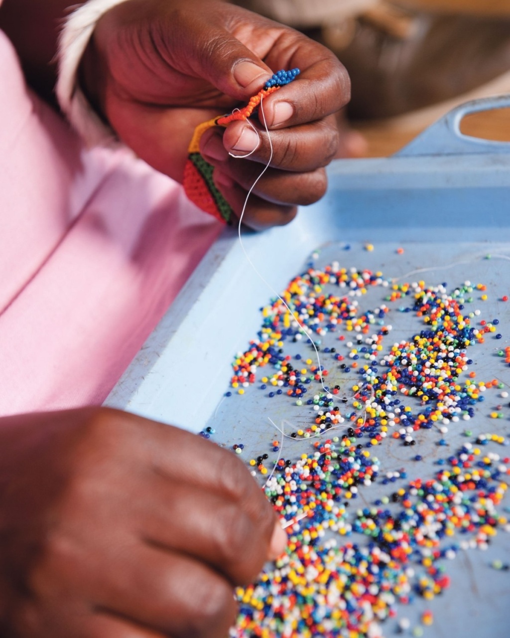 A close-up of hands threading colorful beads on a blue tray. The focus is on the intricate, delicate craft, conveying a sense of creativity and focus.