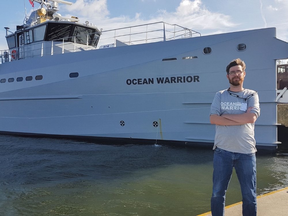 Man stands confidently with arms crossed in front of a large gray ship named "Ocean Warrior." The scene is calm, with a partly cloudy sky.