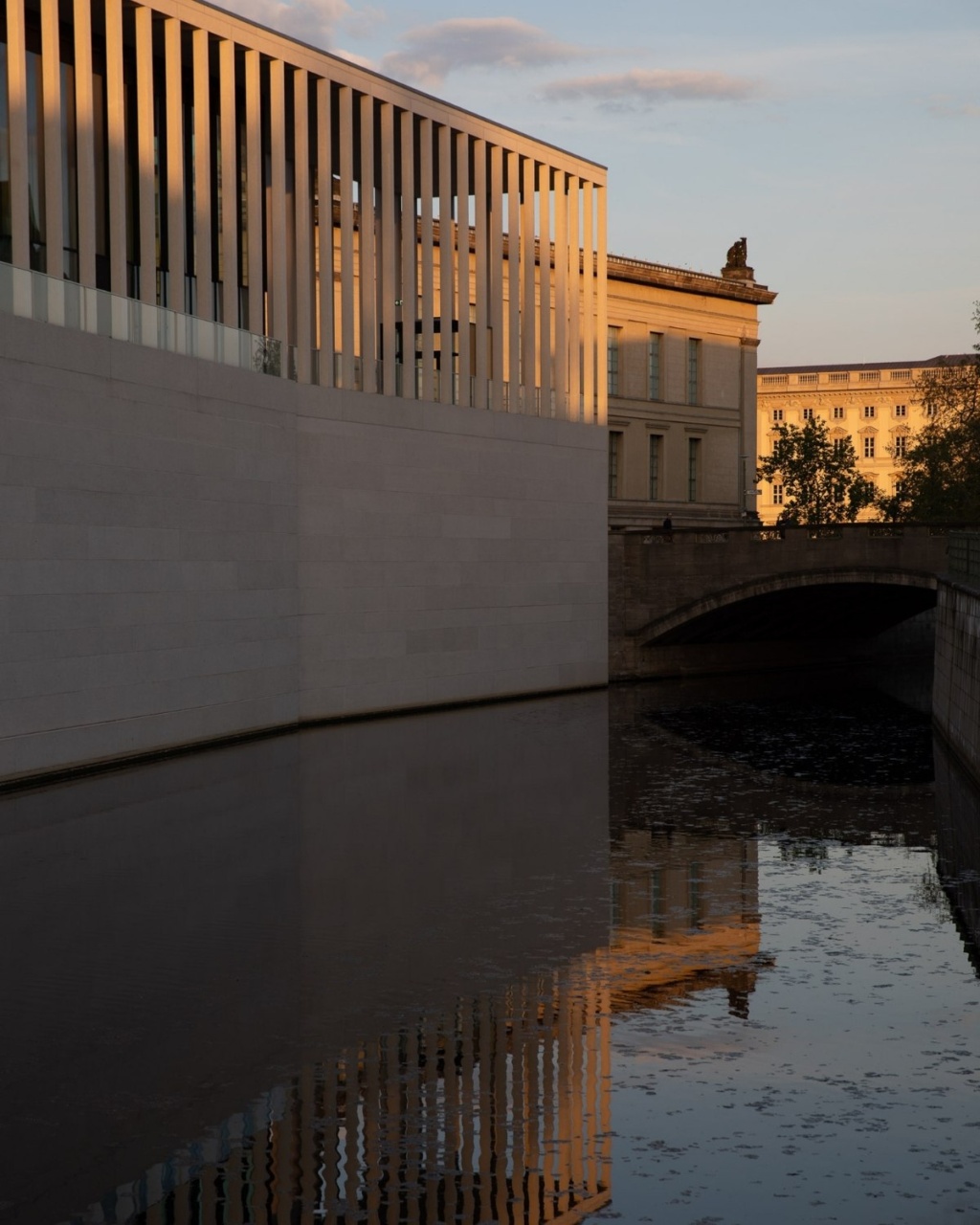 Modern building with vertical windows reflecting on a calm river at sunset. Warm light enhances the architectural lines, creating a serene atmosphere.