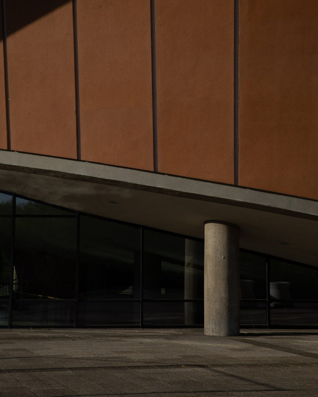 A slanted, modern building facade with terracotta panels and glass windows. A single concrete column provides support, creating a minimalist and industrial feel.