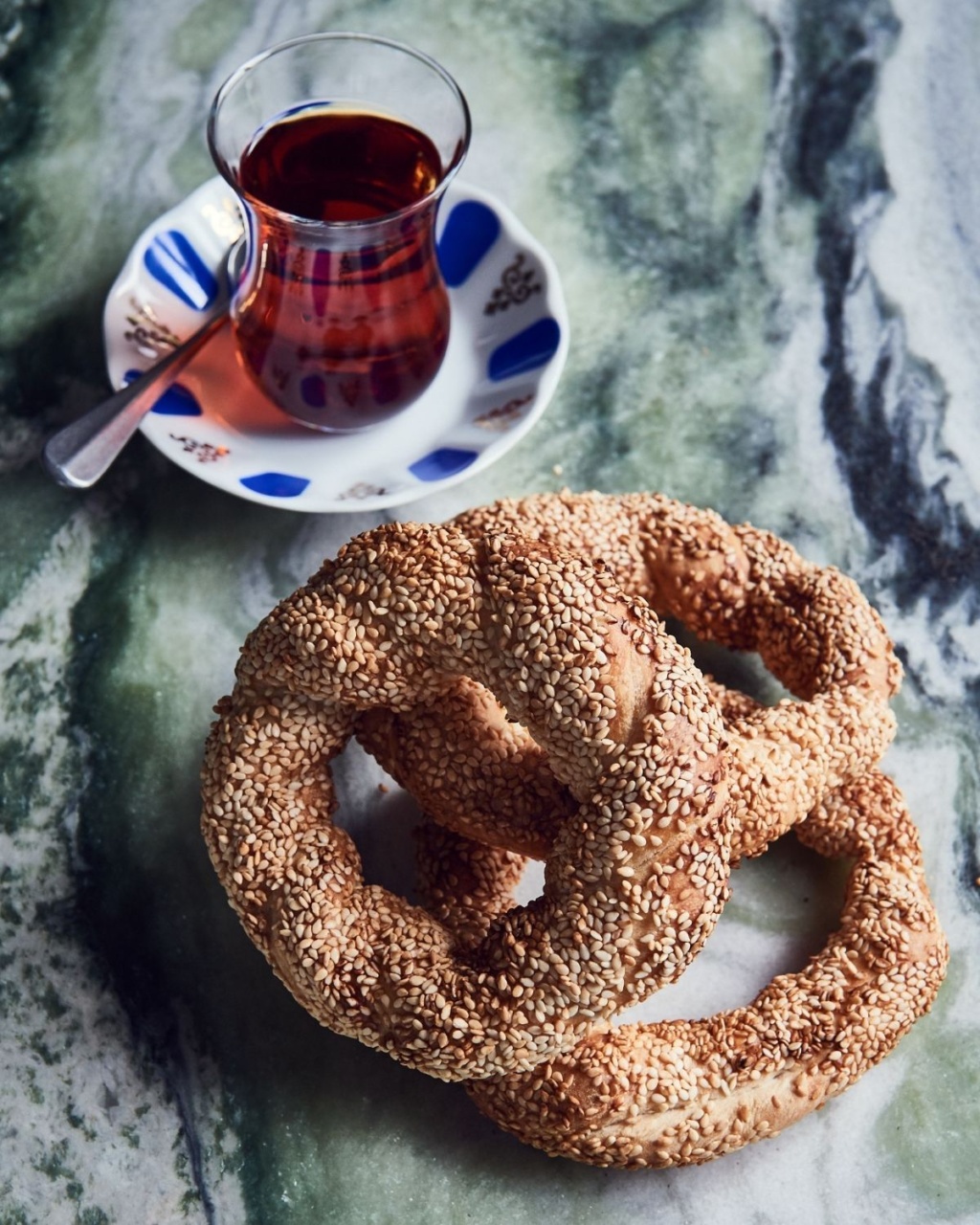 A glass of Turkish tea on a saucer with a spoon beside two sesame-covered simit rings on a marble surface, evoking a cozy and inviting atmosphere.
