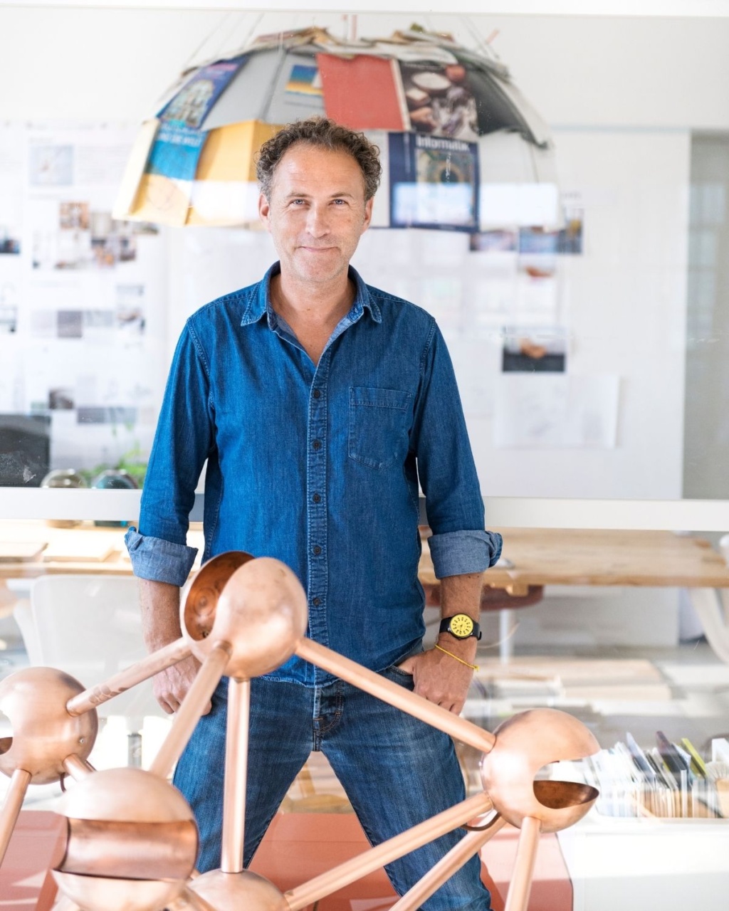 A man in a denim shirt stands confidently in a bright room. A geometric sculpture is in the foreground.