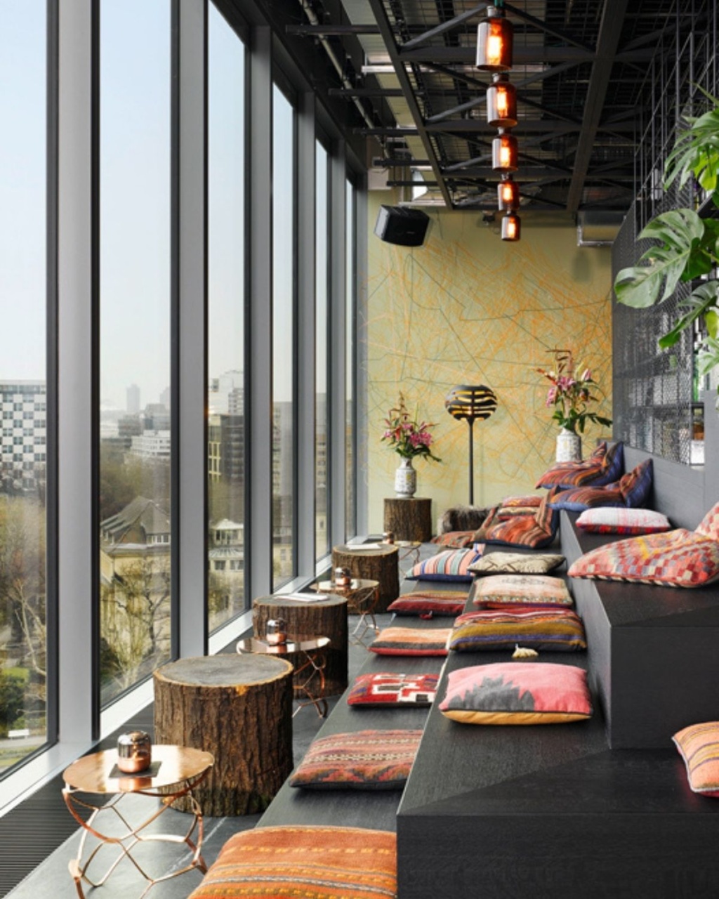 Stylish lounge with tiered seating, vibrant cushions, and tree-stump tables. Large windows offer a city view.