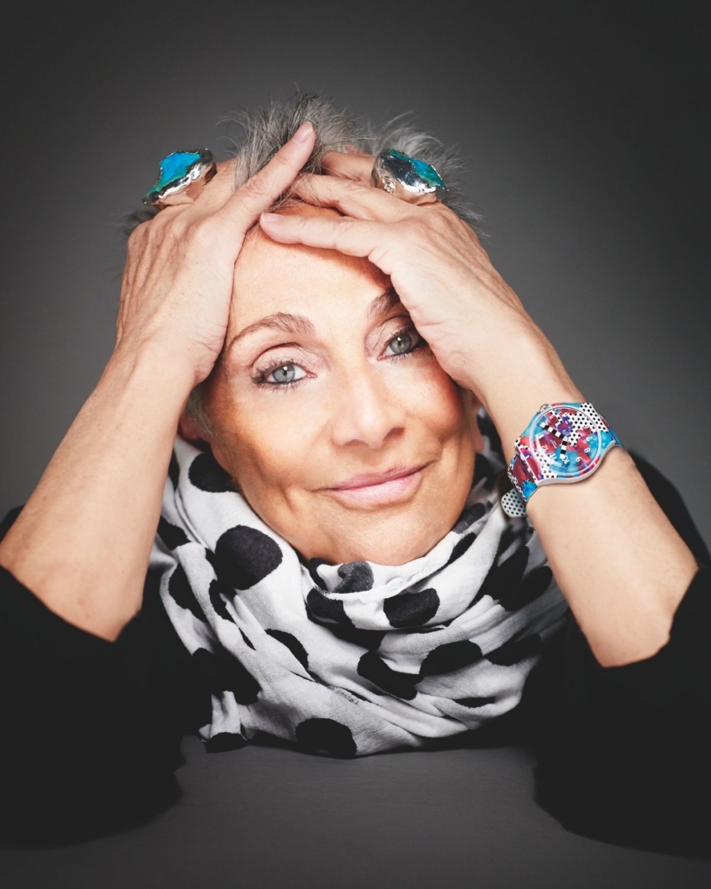 Elderly woman smiles warmly, resting her head in her hands. She wears a black and white polka dot scarf, vibrant jewelry.