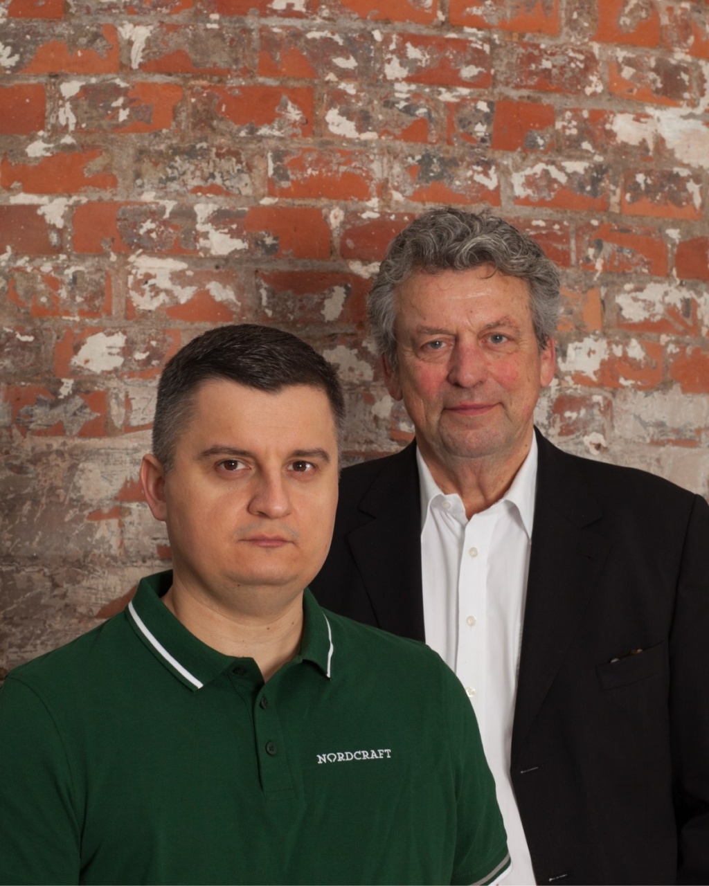 Two men stand side by side against a weathered brick wall. The man on the left wears a green polo shirt, and the man on the right is in a black suit and white shirt. Both appear serious and focused.
