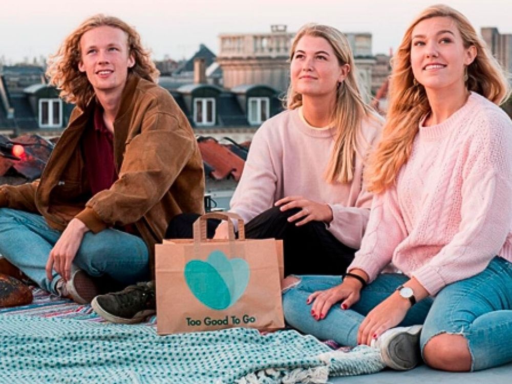 Three young adults sit on a rooftop with relaxed expressions, surrounded by city buildings. A "Too Good To Go" paper bag sits in front of them.