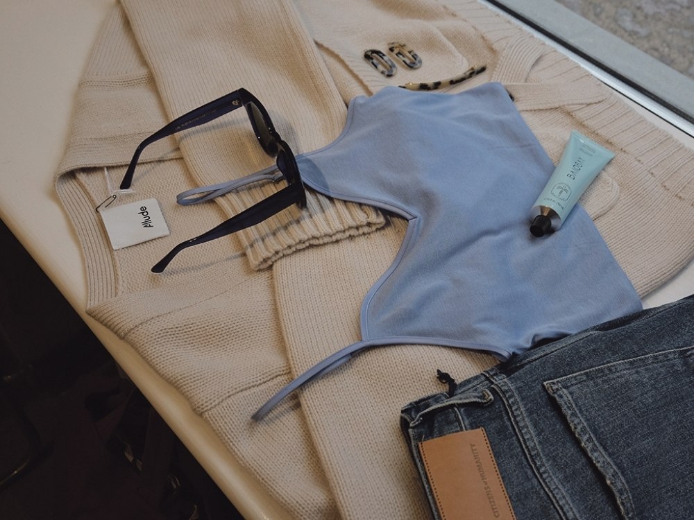Flat lay of a beige sweater, blue top, sunglasses, and hand cream on a table, evoking a chic, casual vibe. Denim jeans are visible on the side.