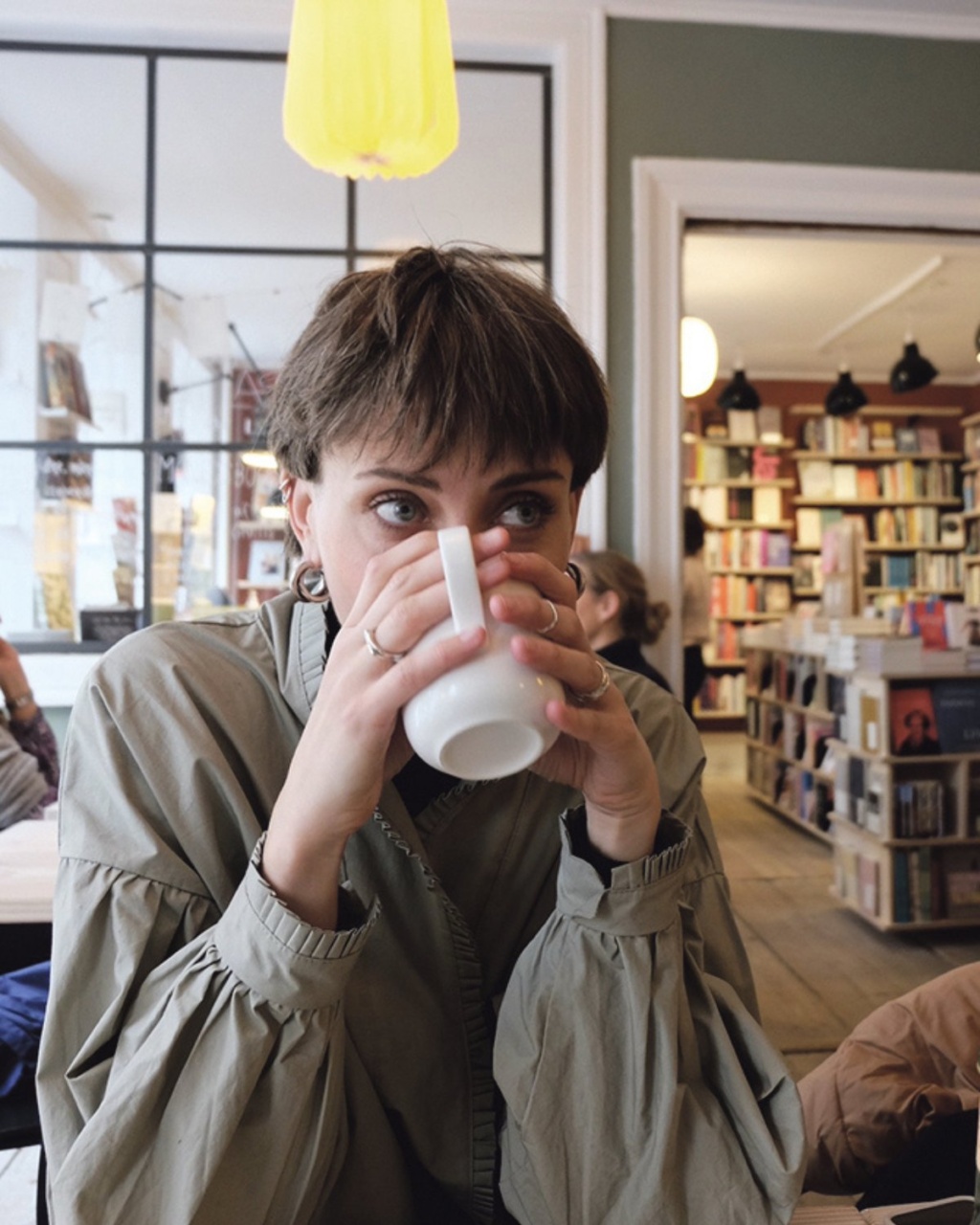 A person with short hair sits in a cozy cafe, sipping from a white mug, surrounded by shelves of books. The atmosphere is warm and inviting.