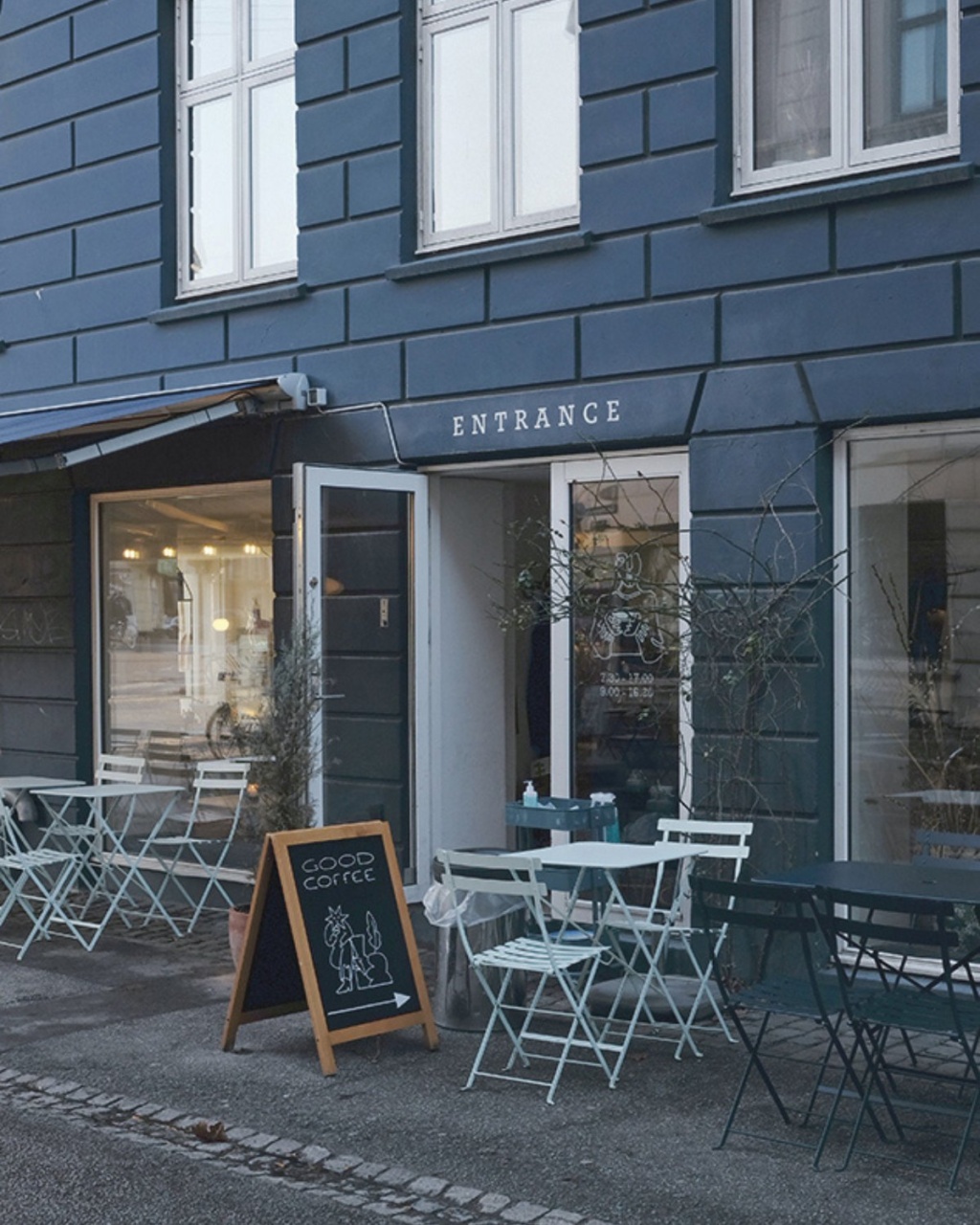 Cozy café exterior with open door, white tables, and chairs set on a quiet street. A chalkboard sign reads "Good Coffee," creating a welcoming vibe.