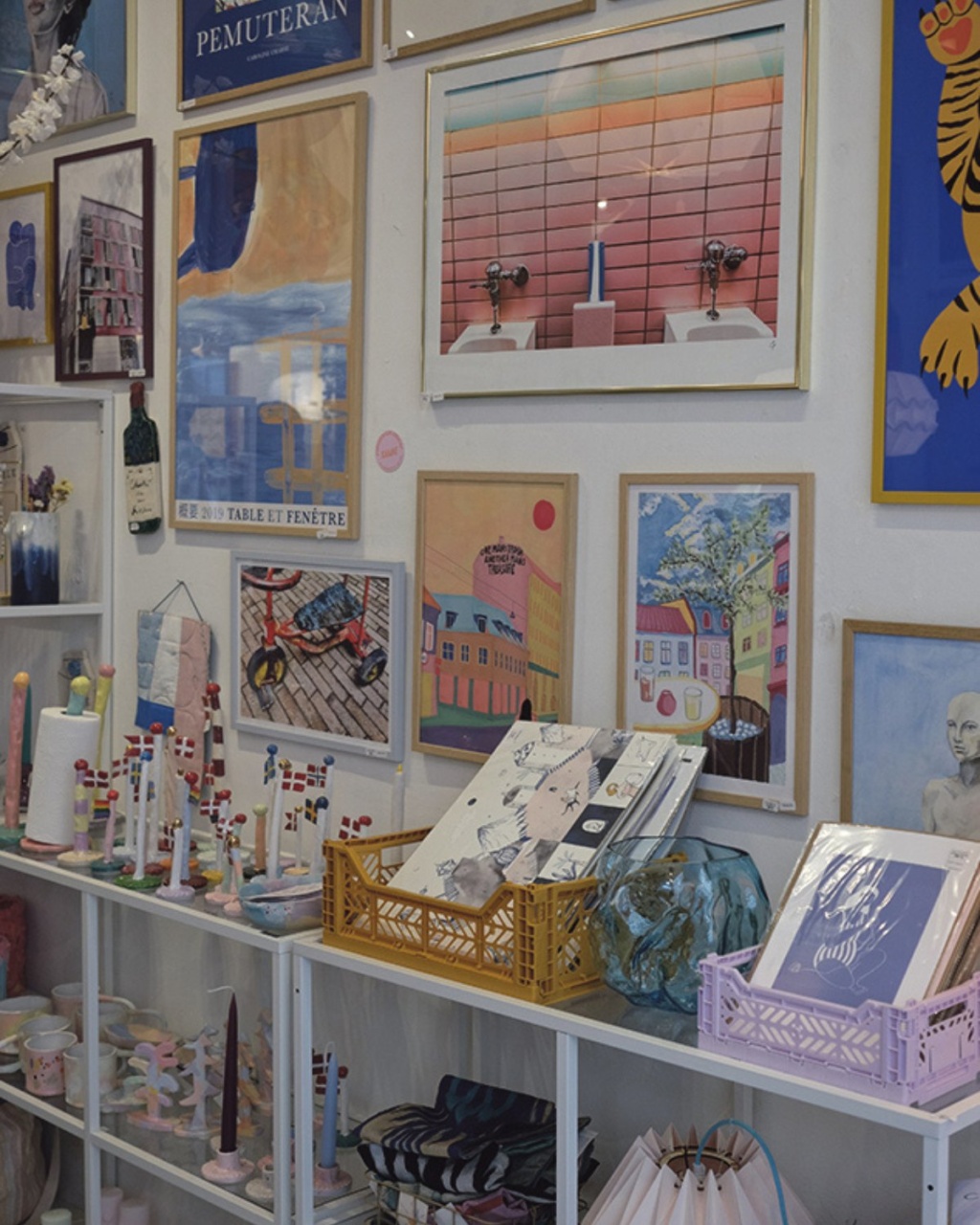 Art gallery wall with colorful framed paintings depicting landscapes, buildings, and abstract designs. Below, shelves display decorative ceramics and candles.