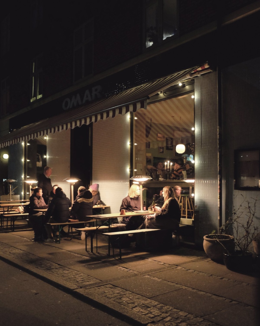 People sit at outdoor tables under soft lighting at a cozy, dimly lit café. Warm, inviting atmosphere with a visible interior showcasing diners.