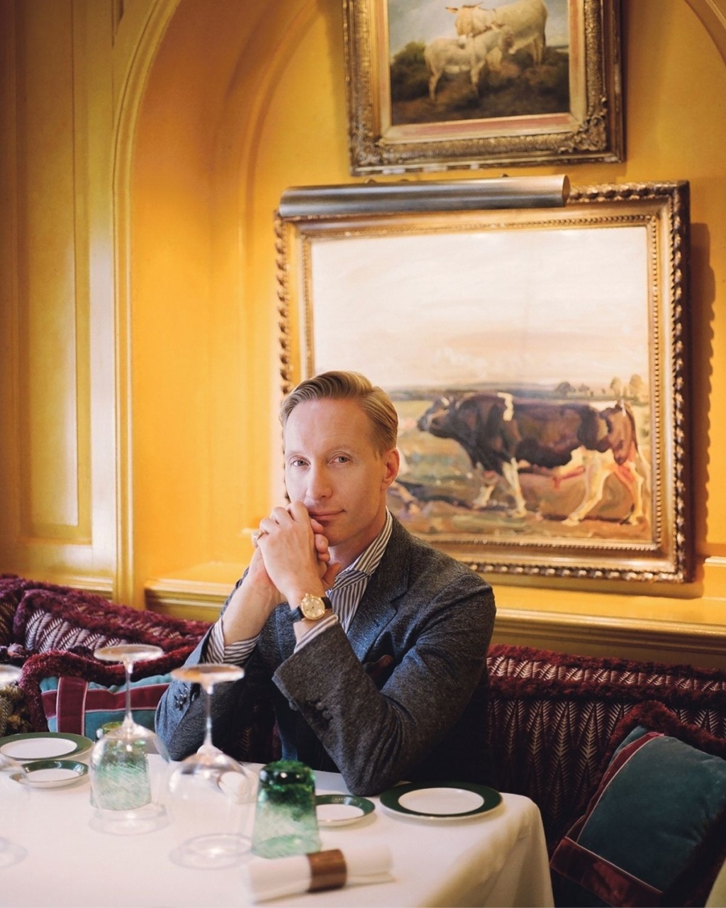 A seated man in formal attire clasps his hands at a dining table with green glassware, against a golden wall adorned with pastoral cow paintings.