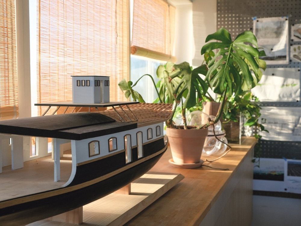 Model ship on wooden table by sunlit window, surrounded by potted plants. Warm tones and soft light create a tranquil, creative atmosphere.