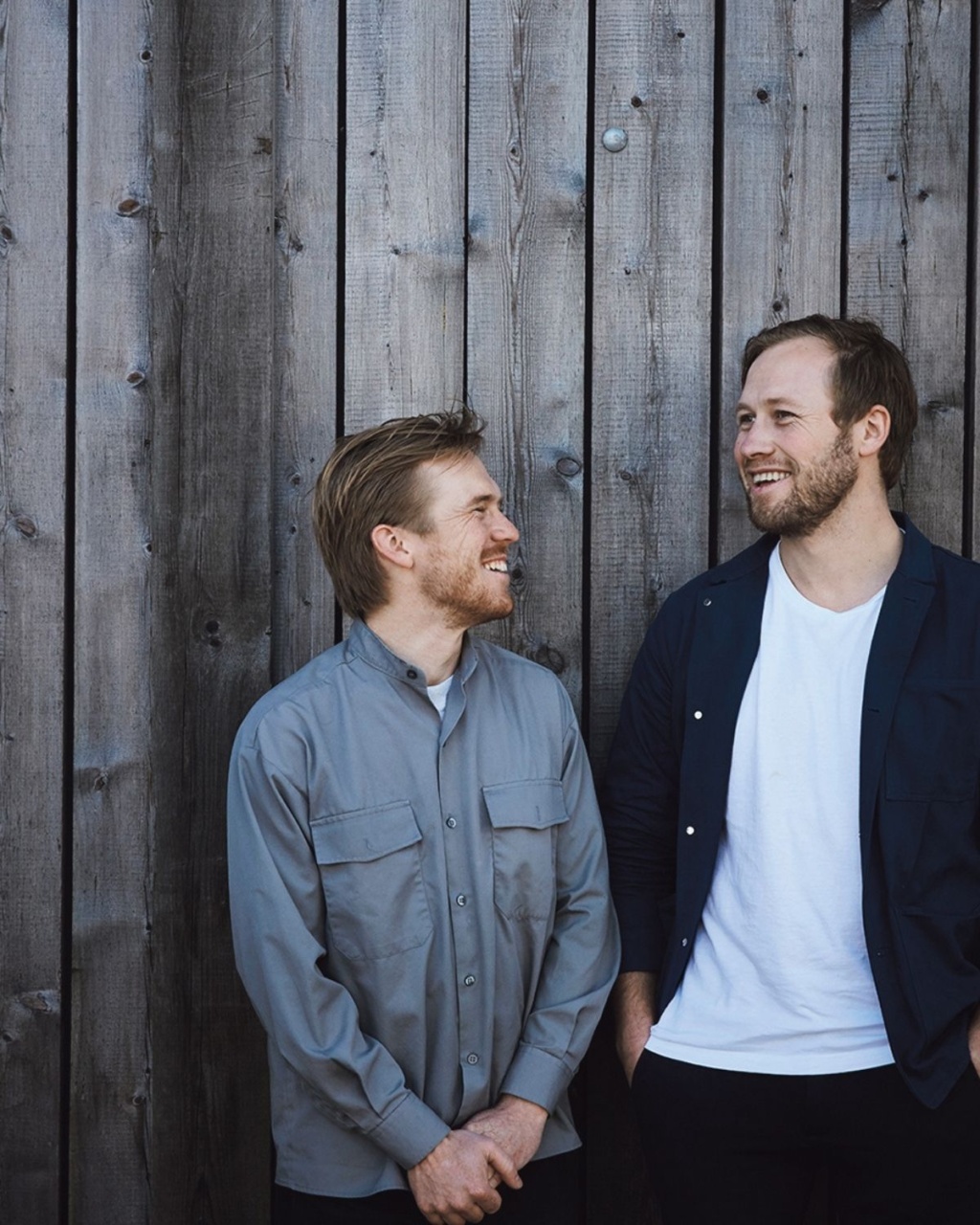 Two men smiling and looking at each other stand against a wooden wall. One wears a gray shirt, the other a navy jacket and white shirt, conveying warmth and friendship.