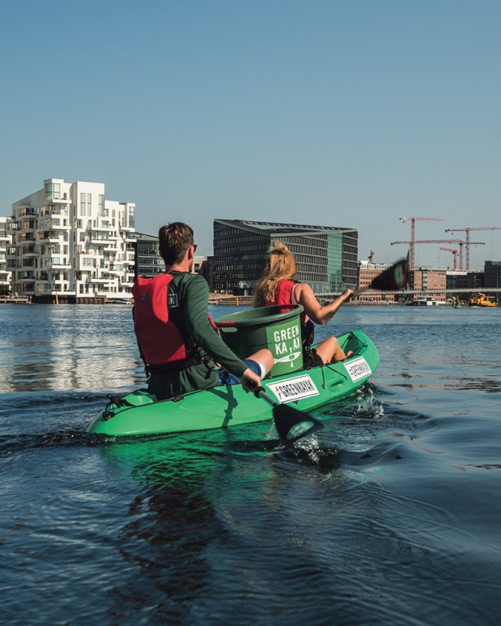 Two people in red life vests paddle a green kayak on calm water, with modern city buildings and cranes in the background under a clear blue sky.