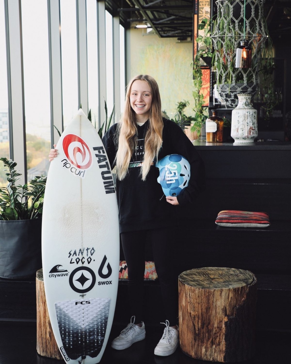Young person with long hair smiling while holding a surfboard and blue helmet indoors. Surrounded by plants, rustic decor, and large windows. Relaxed, sporty vibe.