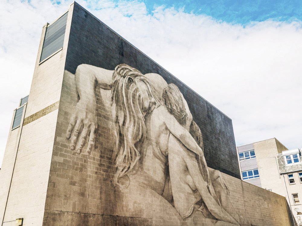 A large mural on a building depicts an embracing couple with long hair, blending shades of gray. The sky is partly cloudy, creating a serene atmosphere.