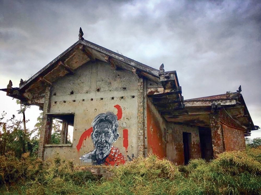 A weathered, abandoned building stands under a cloudy sky. A striking mural of an elderly person is painted on its wall, combining red and grayscale tones. The scene feels somber and evocative.