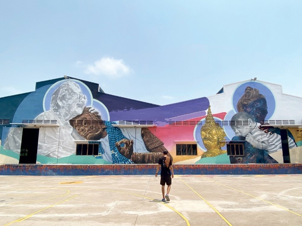 A large mural painted on a warehouse wall portrays figures and a golden Buddha with vibrant colors. A person in black walks toward it, evoking reflection.
