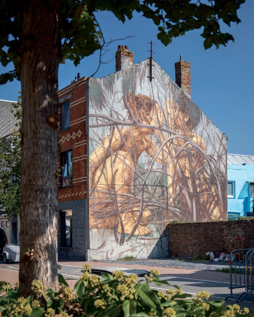 Large mural of intertwined branches covers the side of a brick building, creating an organic, textured pattern. Framed by a tree and plants, under a clear sky.