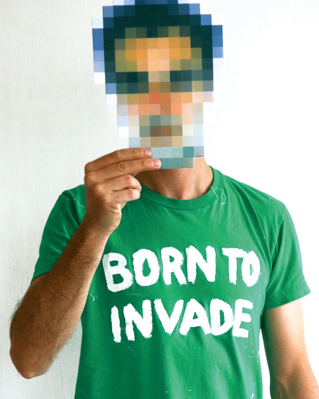 Man in a green shirt with "BORN TO INVADE" text holds a pixelated image over his face. The background is plain white, creating a humorous tone.