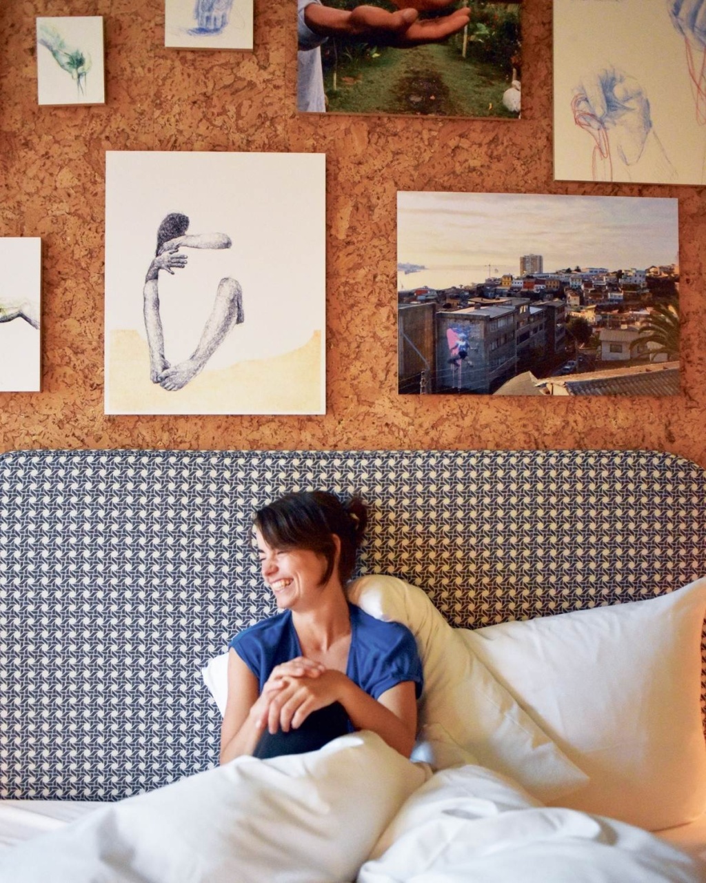 A woman sits in bed with a joyful expression, surrounded by artwork on a cork wall. The room feels cozy, blending restful and artistic elements.