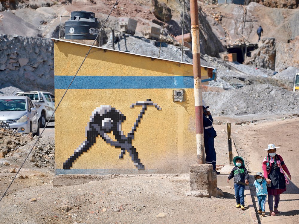 A yellow building with a pixel art miner holding a pickaxe. Nearby, a woman and two children in masks walk on a dirt road. The area is rocky and rugged.