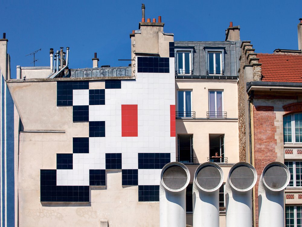Building facade with mosaic art of a pixelated character made of black, white, and red tiles. Four large white vents are in front. Clear blue sky.