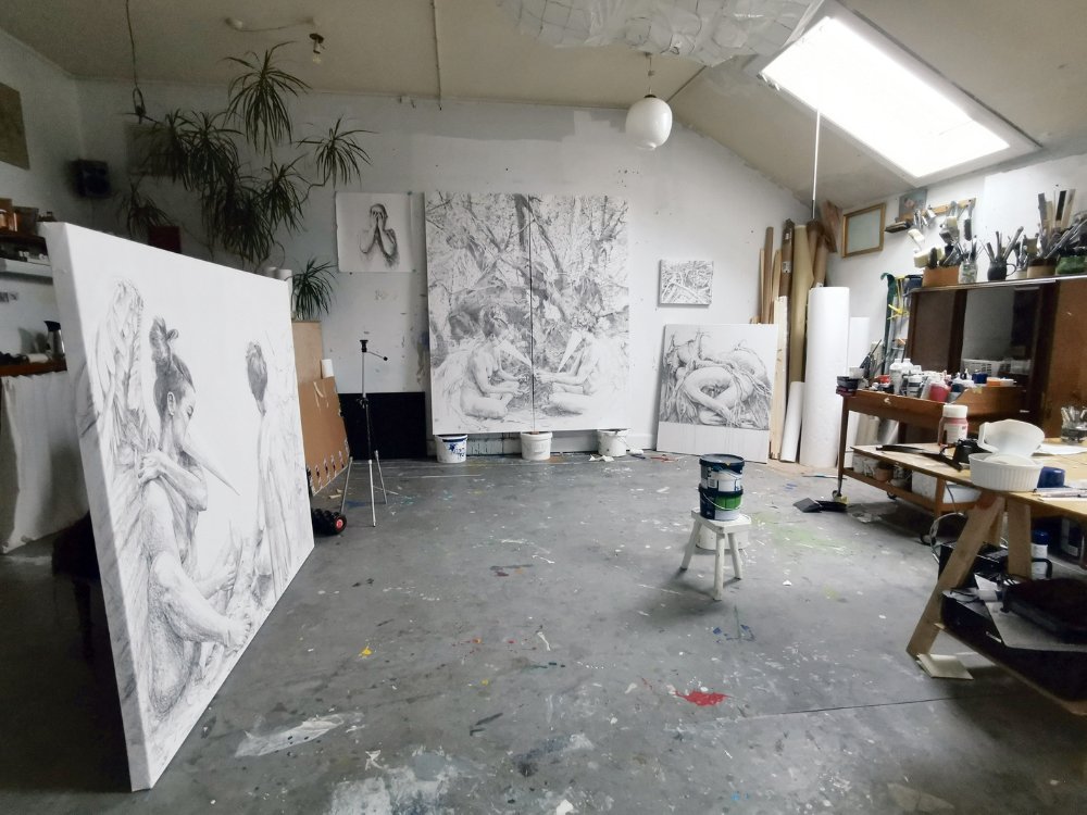 Art studio with large monochrome paintings leaning against the walls, depicting serene, contemplative figures. A cluttered workspace with paint supplies fills the room. Soft lighting from skylight creates an artistic and focused atmosphere.